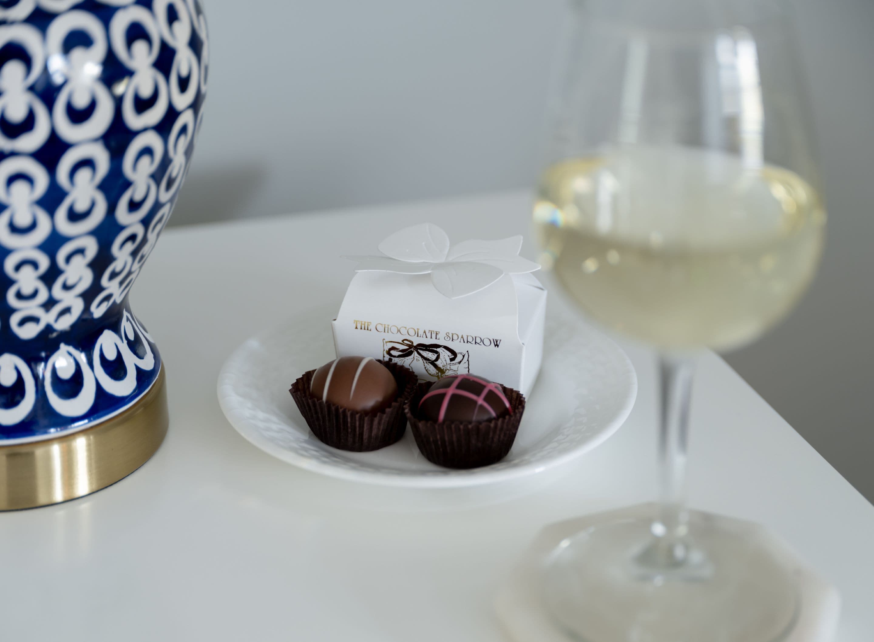 A white table with a blue and gold vase, a glass of white wine, and a box labeled "The Chocolate Sparrow" with chocolates.