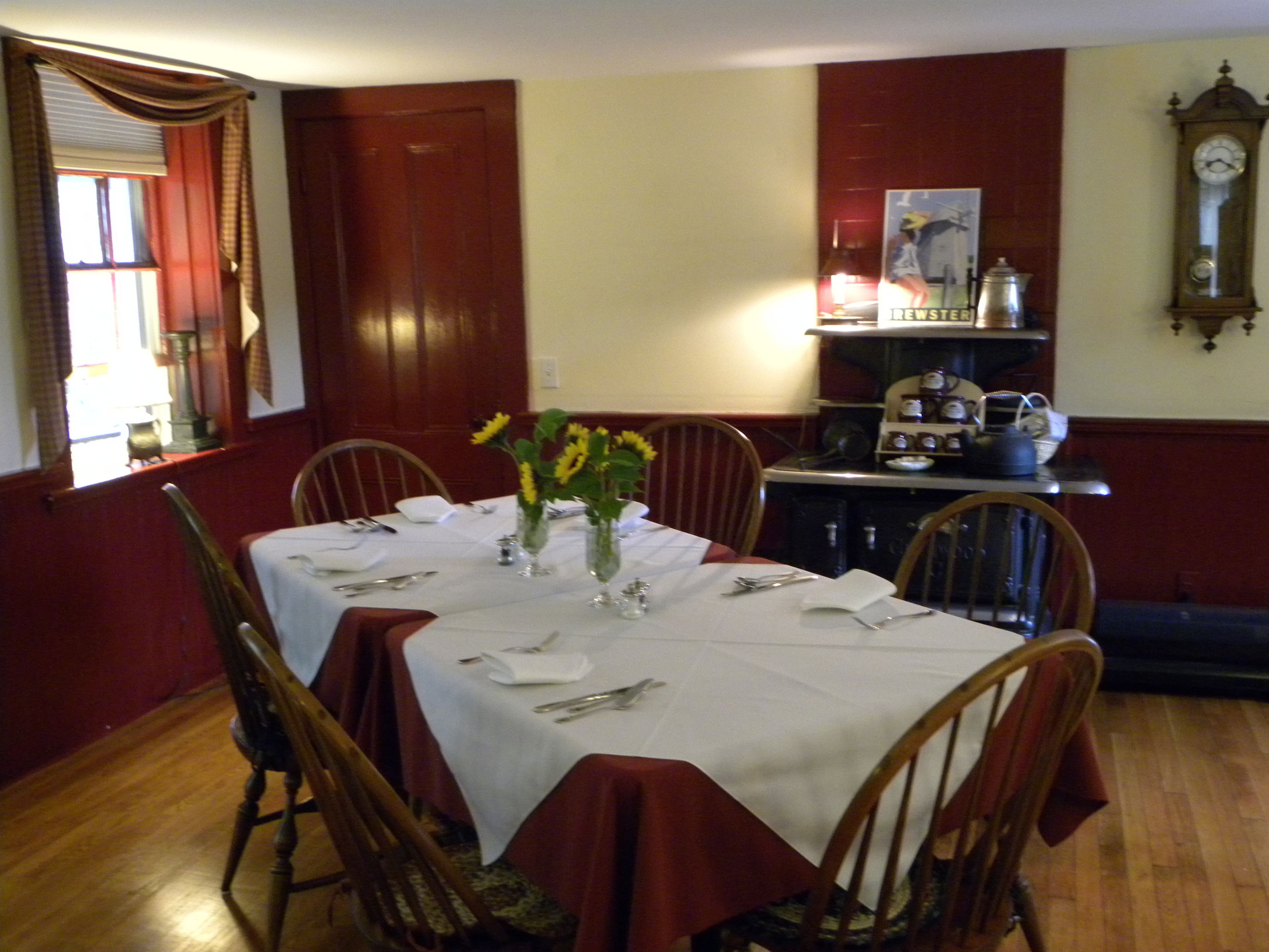 Une salle à manger avec une table dressée pour quatre personnes, entourée de chaises en bois. On peut voir des vases remplis de fleurs sur la table, et une horloge au mur.