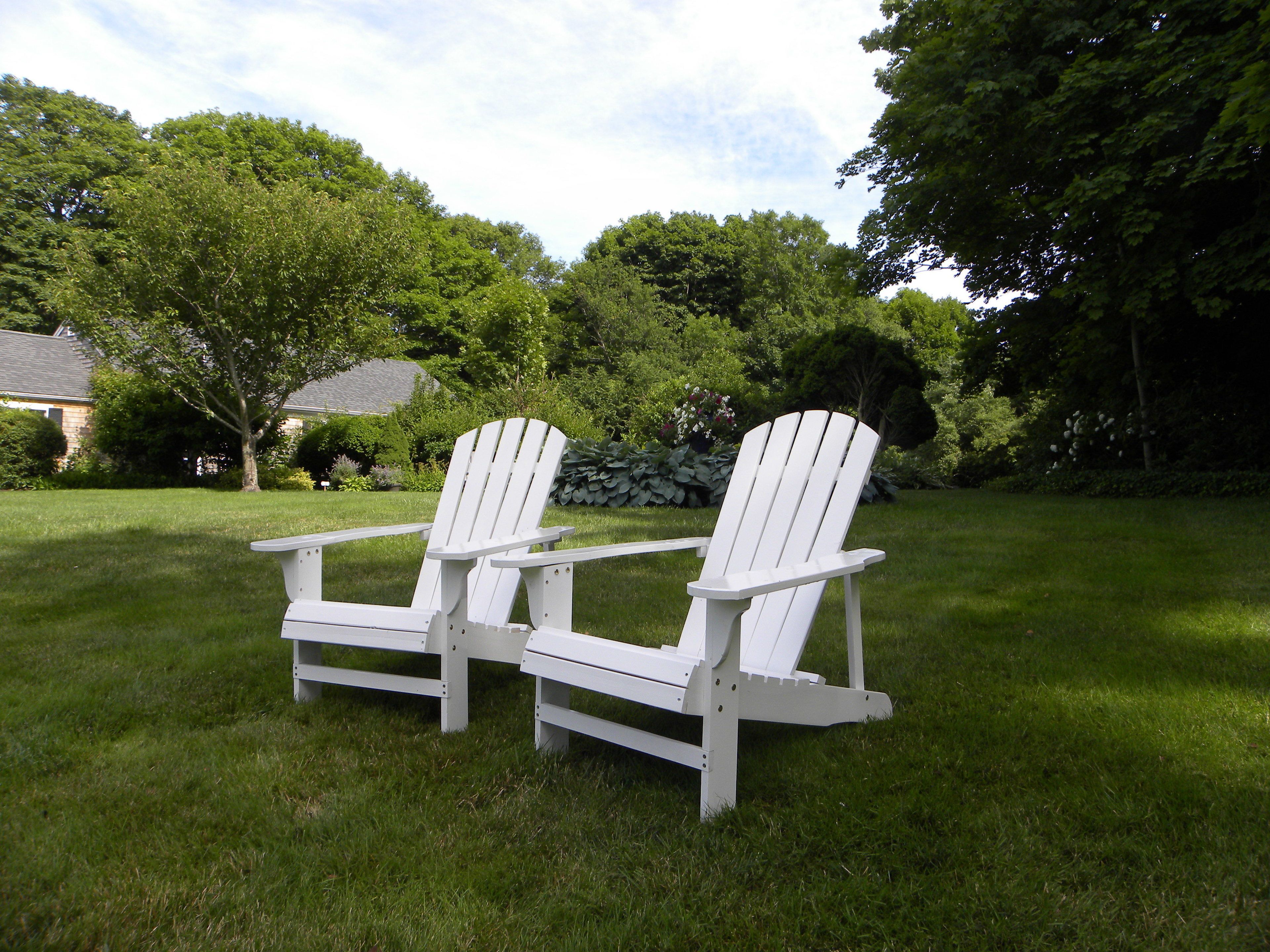 Two white Adirondack chairs in a grassy yard with trees and a building in the background.