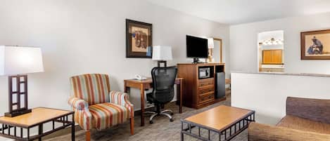 A hotel room with a sofa, armchair, desk, TV, and a kitchenette.