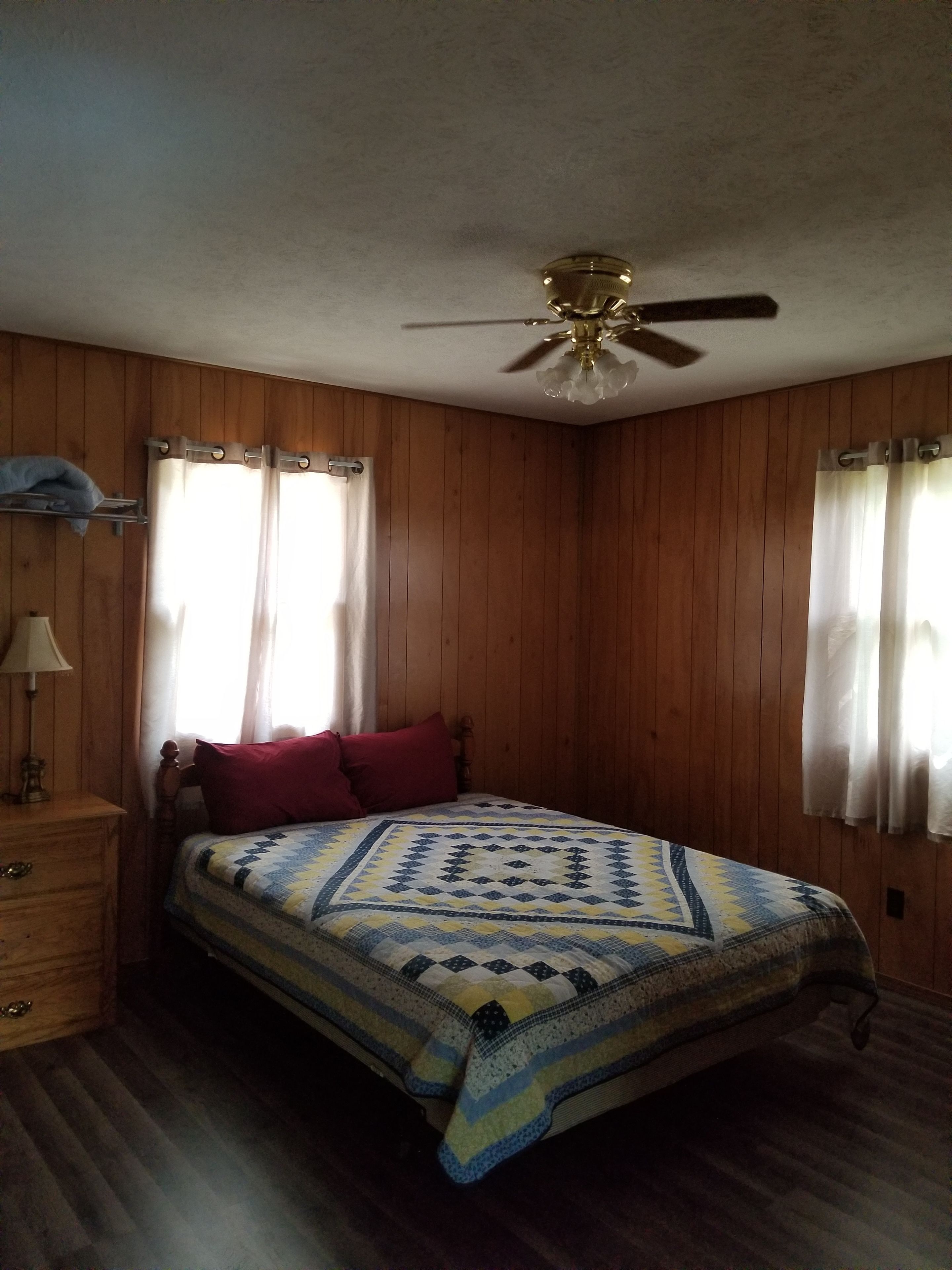 Un dormitorio con paredes de madera, una cama con manta estampada, una mesita de noche con lámpara y un ventilador de techo.