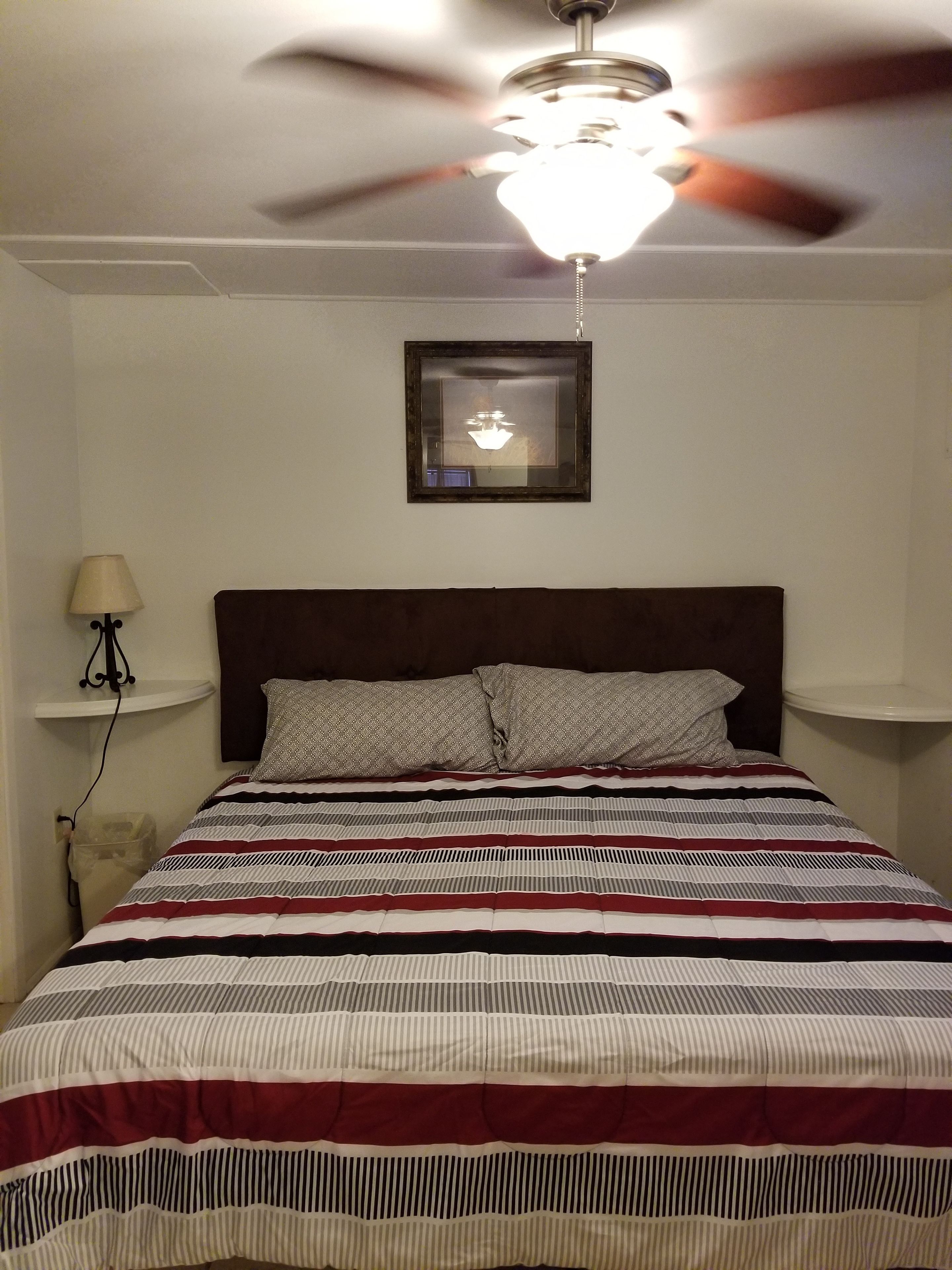 A bedroom with a bed, a ceiling fan, a framed picture on the wall, and a nightstand with a lamp.