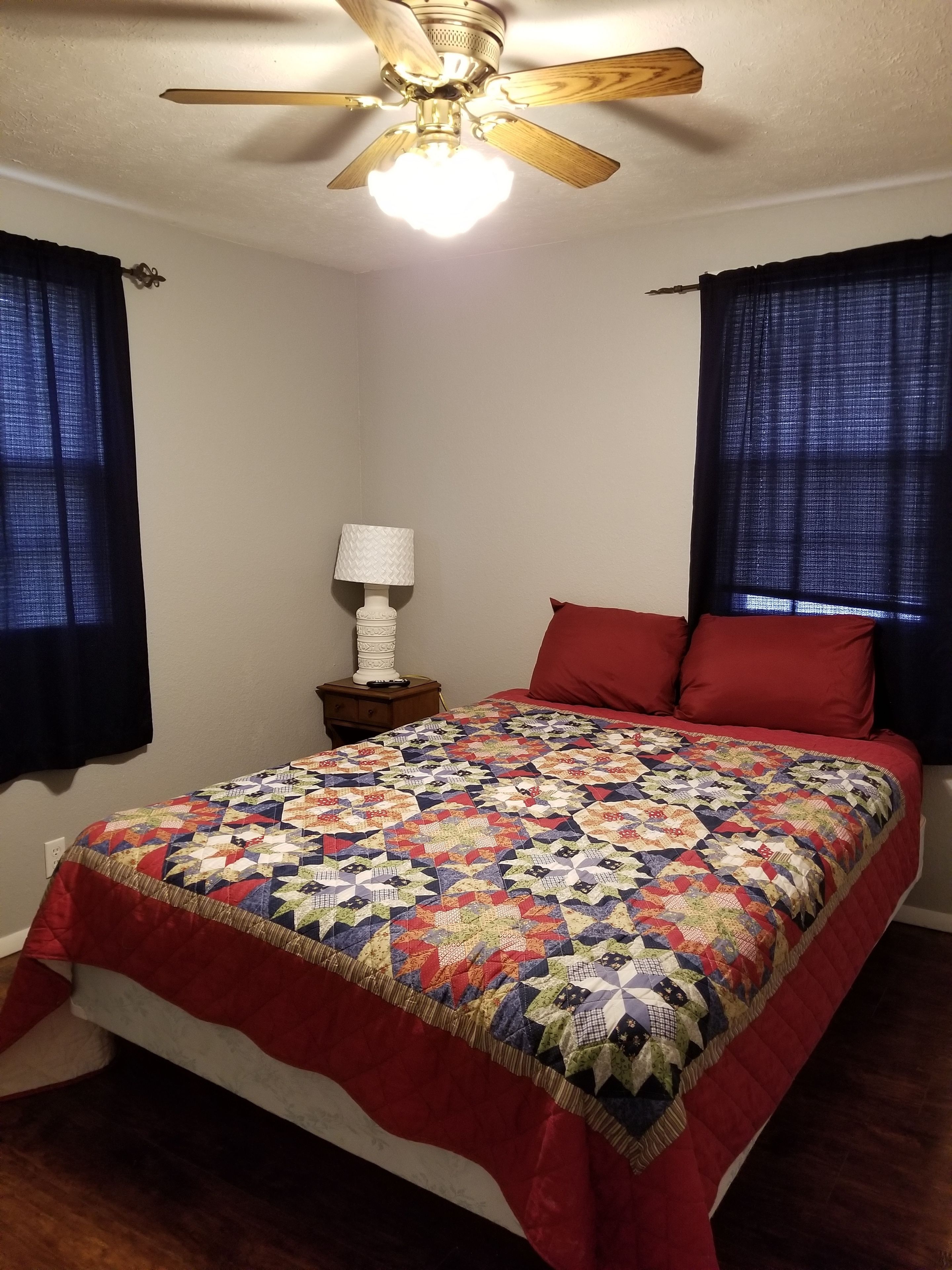 Un dormitorio con ventilador de techo de madera, una cama con colcha de colores, una mesita de noche con lámpara y cortinas oscuras.