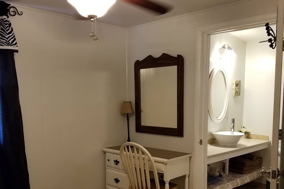 A bedroom with a bed, a desk with a chair, a dresser, a mirror, a ceiling fan, and a bathroom with a sink and toilet.