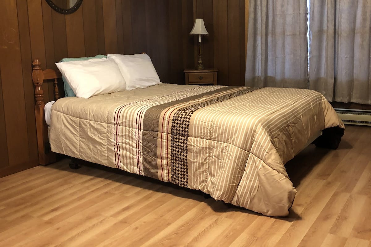 A bedroom with a wooden floor, a bed with a patterned bedspread, a nightstand with a lamp, a round mirror, and a window with curtains.