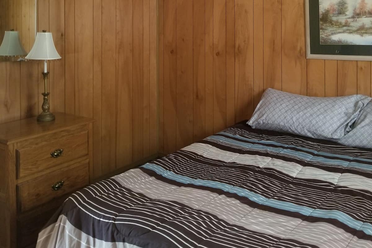 A bedroom with wooden paneling, a bed with striped bedding, a nightstand with a lamp, and a ceiling fan.
