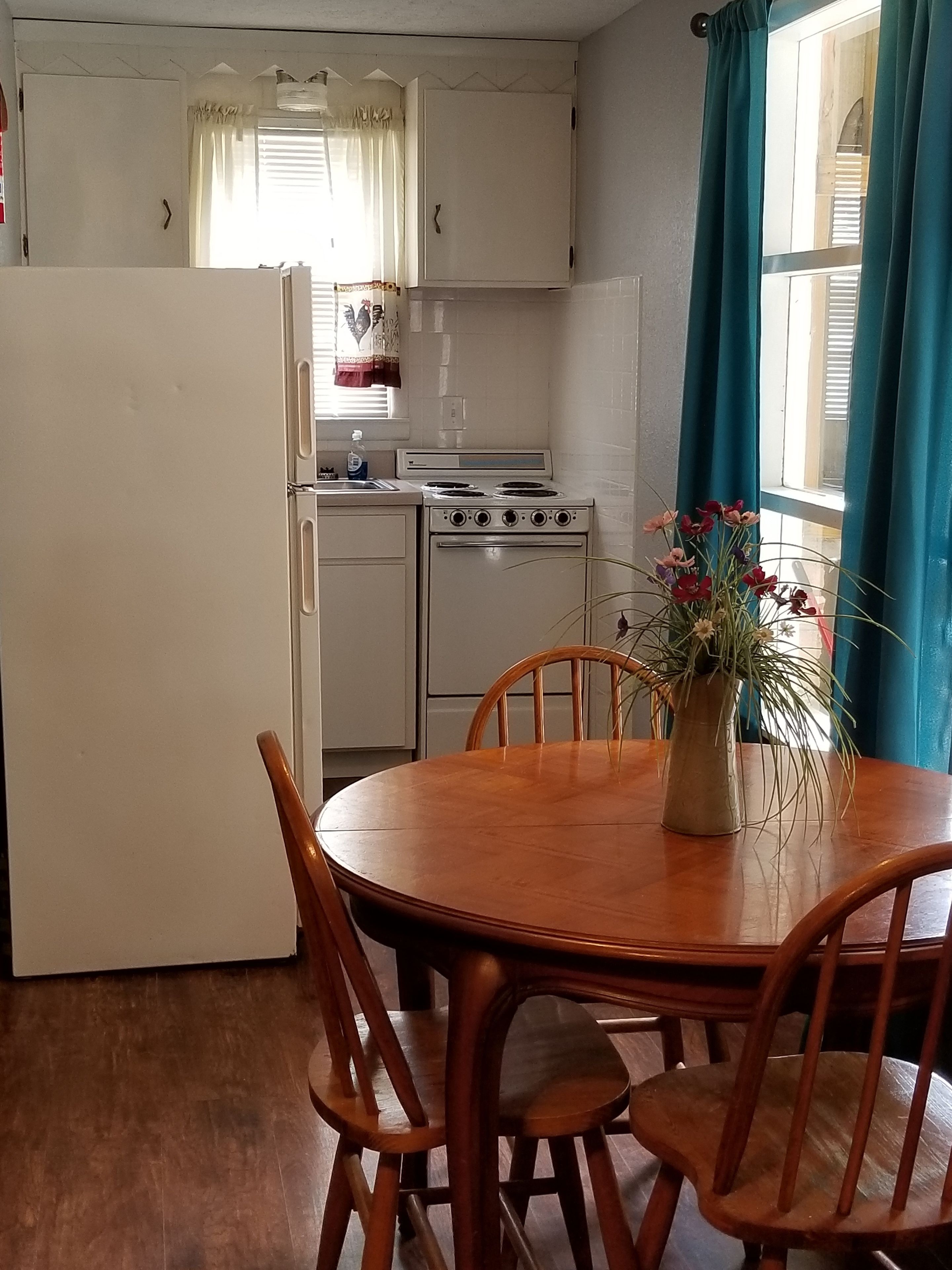 A compact kitchen with a white refrigerator, stove, and sink, a wooden dining table with chairs, and a window with curtains.