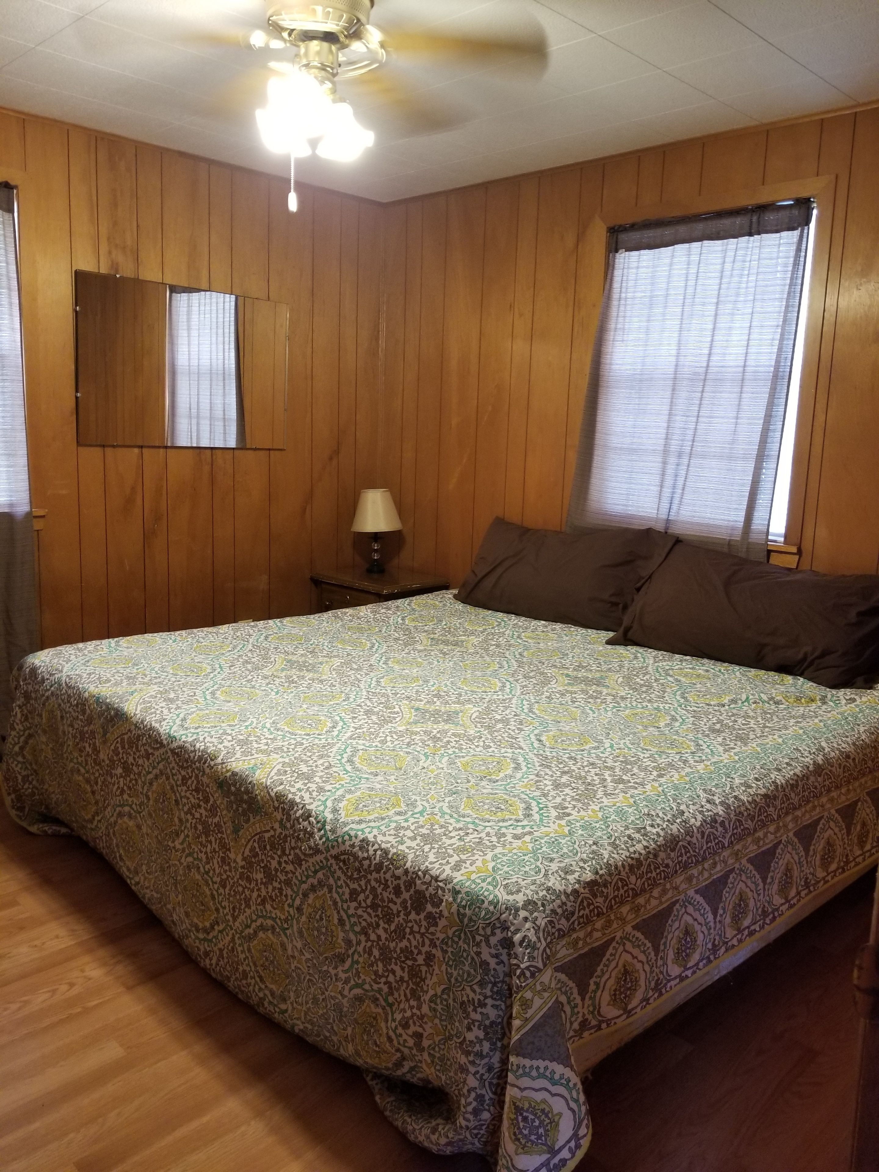 Un dormitorio con pared de paneles de madera, cama con ropa de cama estampada, mesita de noche con lámpara y ventilador de techo.