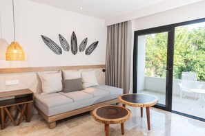 A modern living room with a sofa, two round wooden tables, a wall-mounted light fixture, and a large sliding glass door leading to an outdoor area. - Bahía Príncipe Explore Akumal (Riviera Maya)