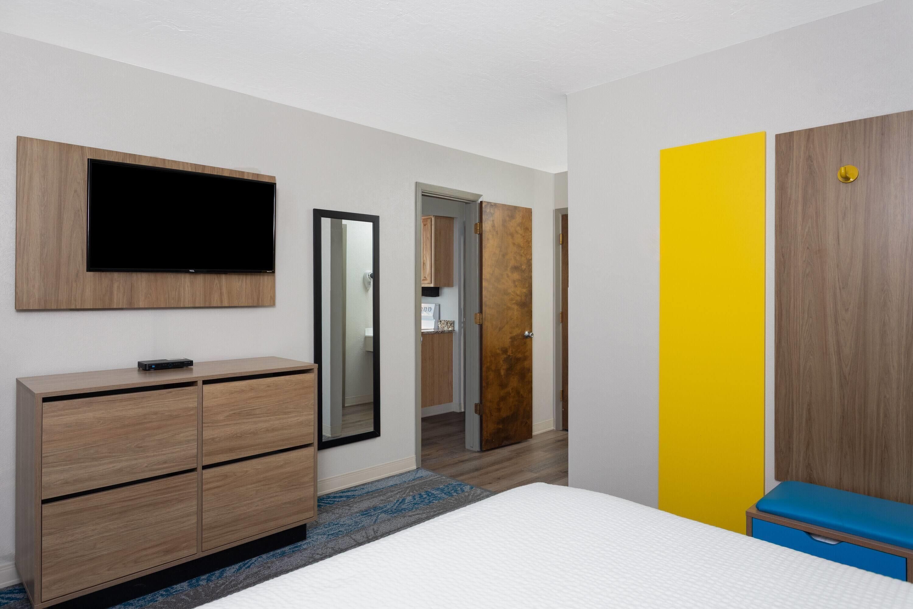 Une chambre d’hôtel moderne dotée d’un téléviseur à écran plat fixé au mur, d’une commode en bois, d’un lit avec un banc bleu et d’un mur d’accent jaune.