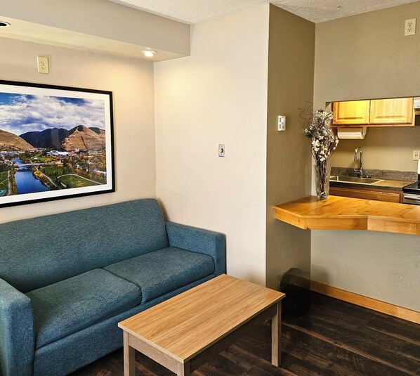 A modern living room with a blue sofa, a small wooden table, and a kitchen area with wooden cabinets and a sink.