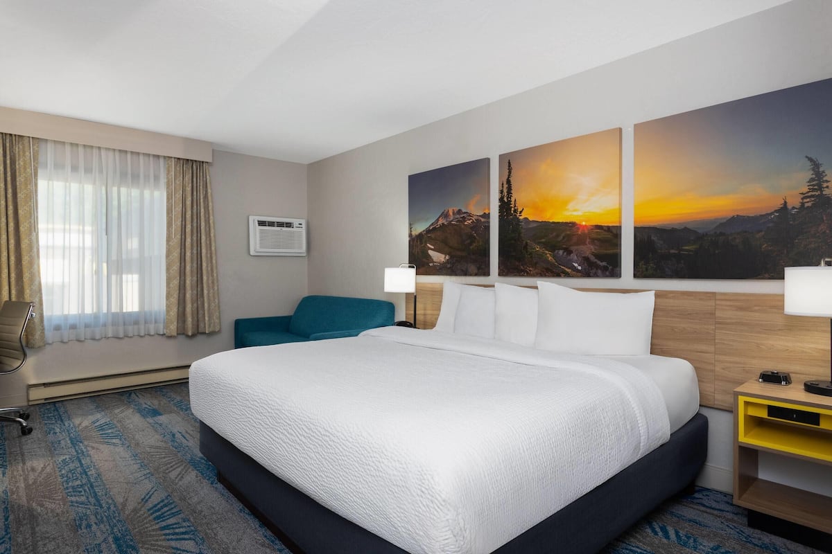 A hotel room with a large bed, a desk with a lamp, a chair, and two paintings on the wall.