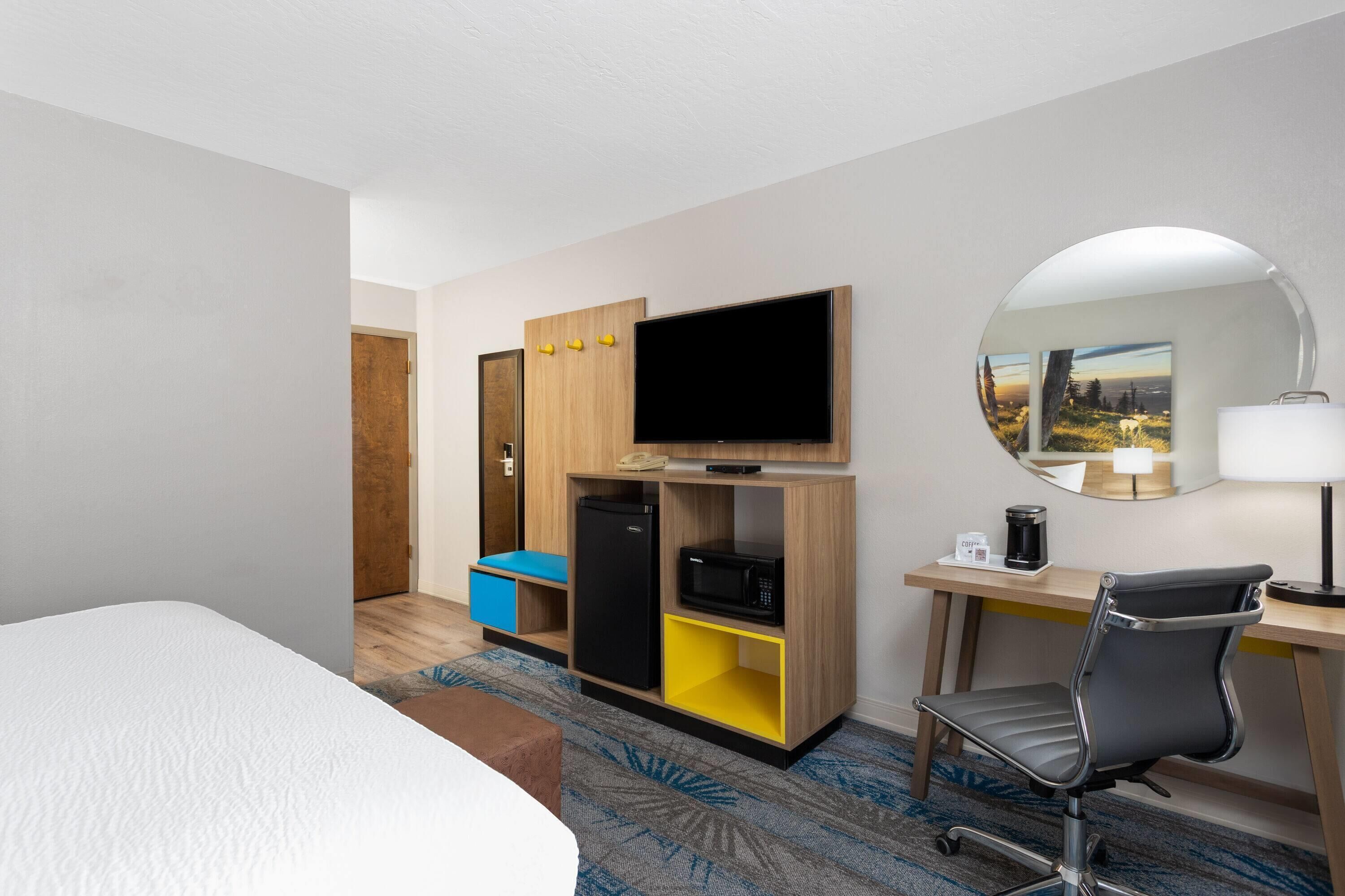 A hotel room with a bed, a desk with a chair, a TV, a mirror, and a minibar.