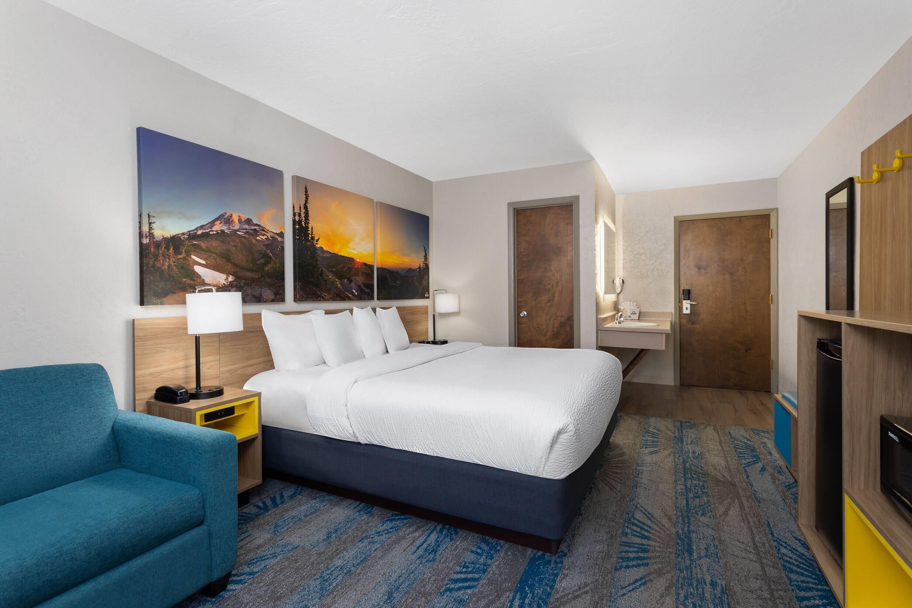 Een hotelkamer met een groot bed, een blauwe fauteuil, een houten nachtkastje, een bureau met spiegel en een schilderij van een berglandschap.