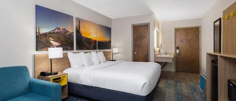 Een hotelkamer met een groot bed, een blauwe fauteuil, een houten nachtkastje, een bureau met spiegel en een schilderij van een berglandschap.