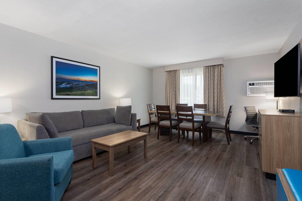A modern hotel room with a grey sofa, a blue armchair, a wooden coffee table, a dining table with chairs, a flat-screen TV, and an air conditioning unit.