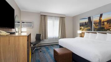 A hotel room with a bed, a desk, a chair, a television, an air conditioning unit, and a window with curtains.