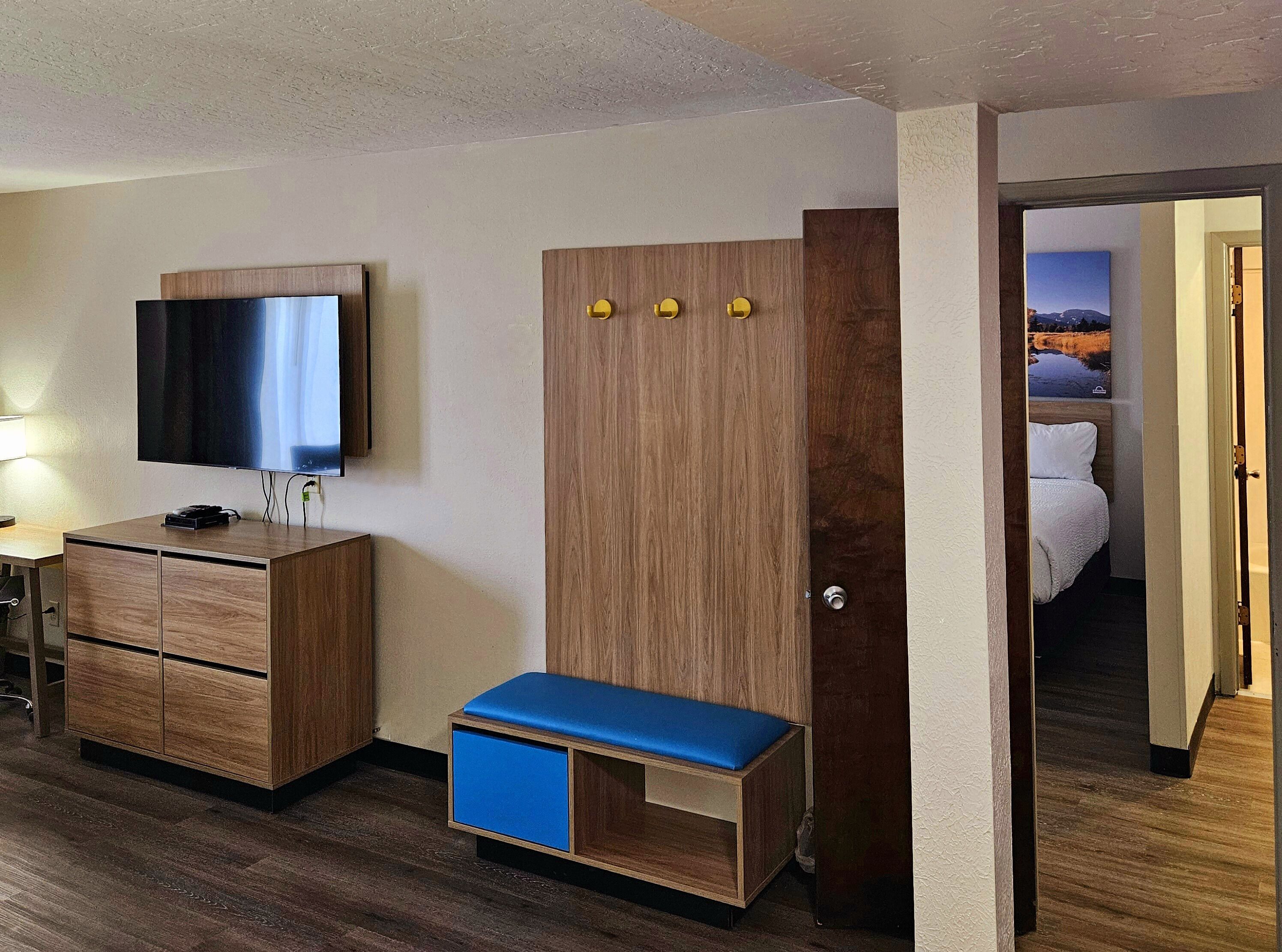 Habitación de hotel con armario de madera, televisor montado en la pared, un banco azul y cama con ropa de cama blanca.