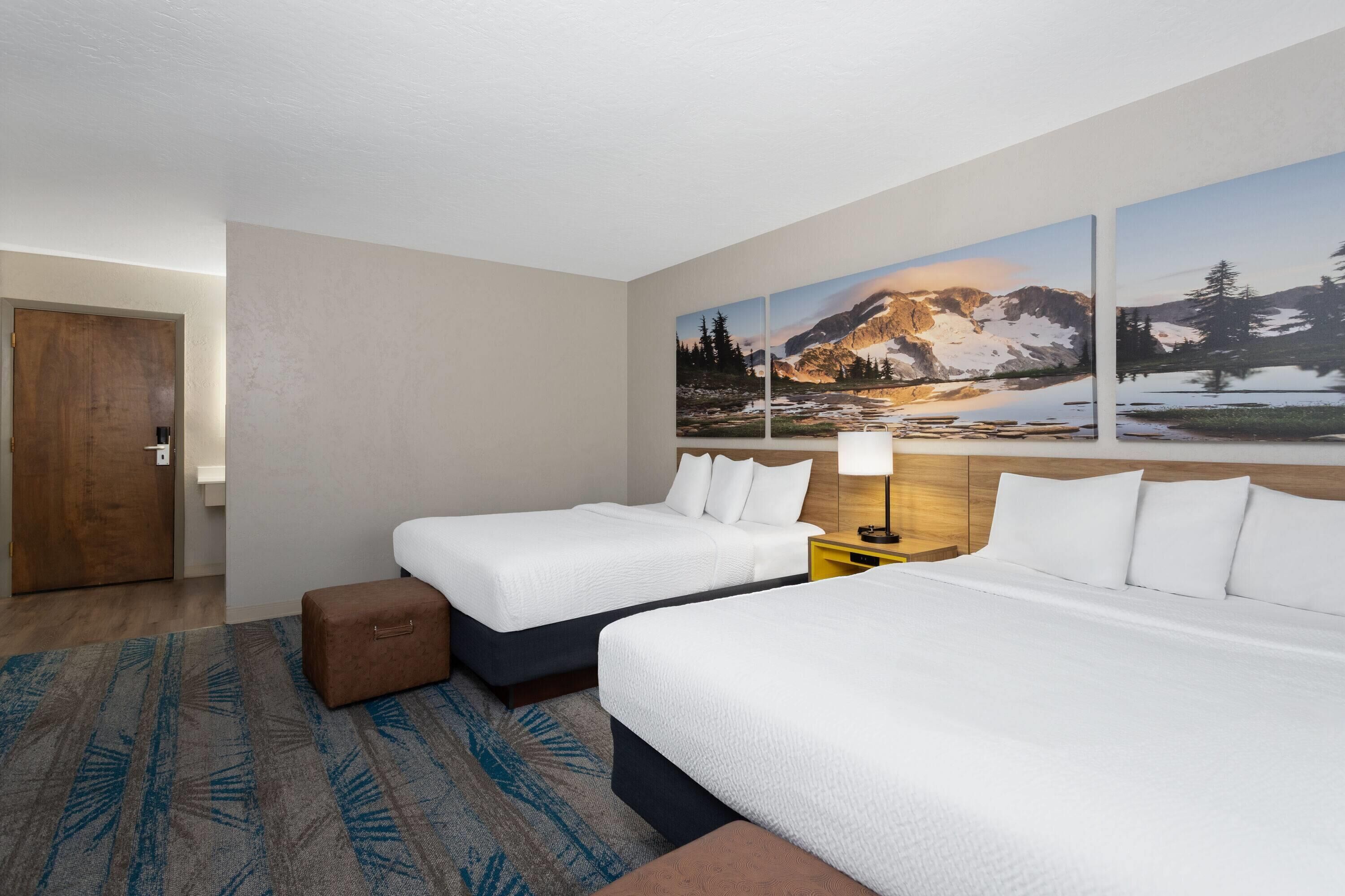 A hotel room with two beds, a wooden door, a nightstand with a lamp, and a large mountain landscape painting on the wall.