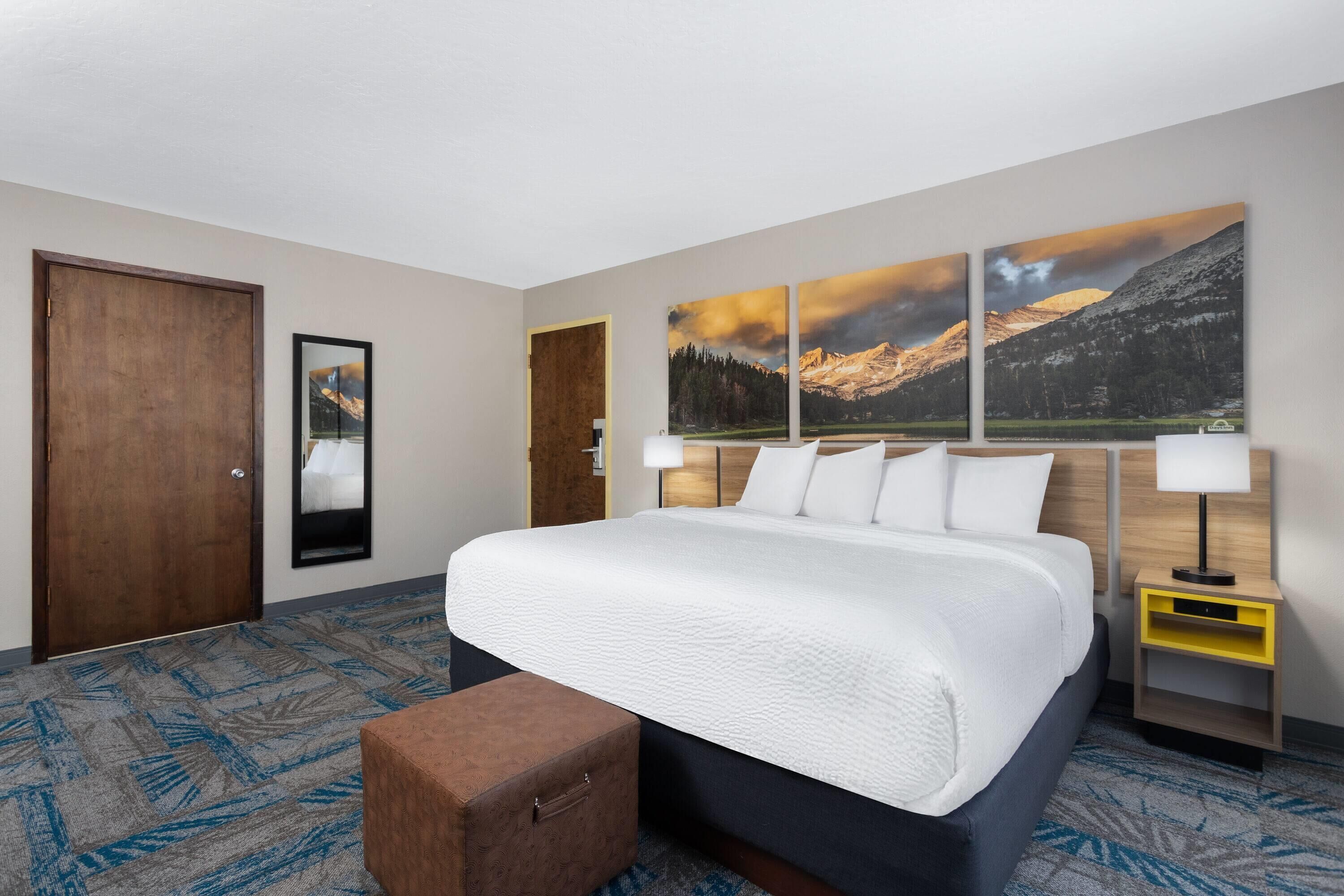 A hotel room with a large bed, a wooden door, a mirror, a nightstand with a lamp, and a wall with three framed pictures.
