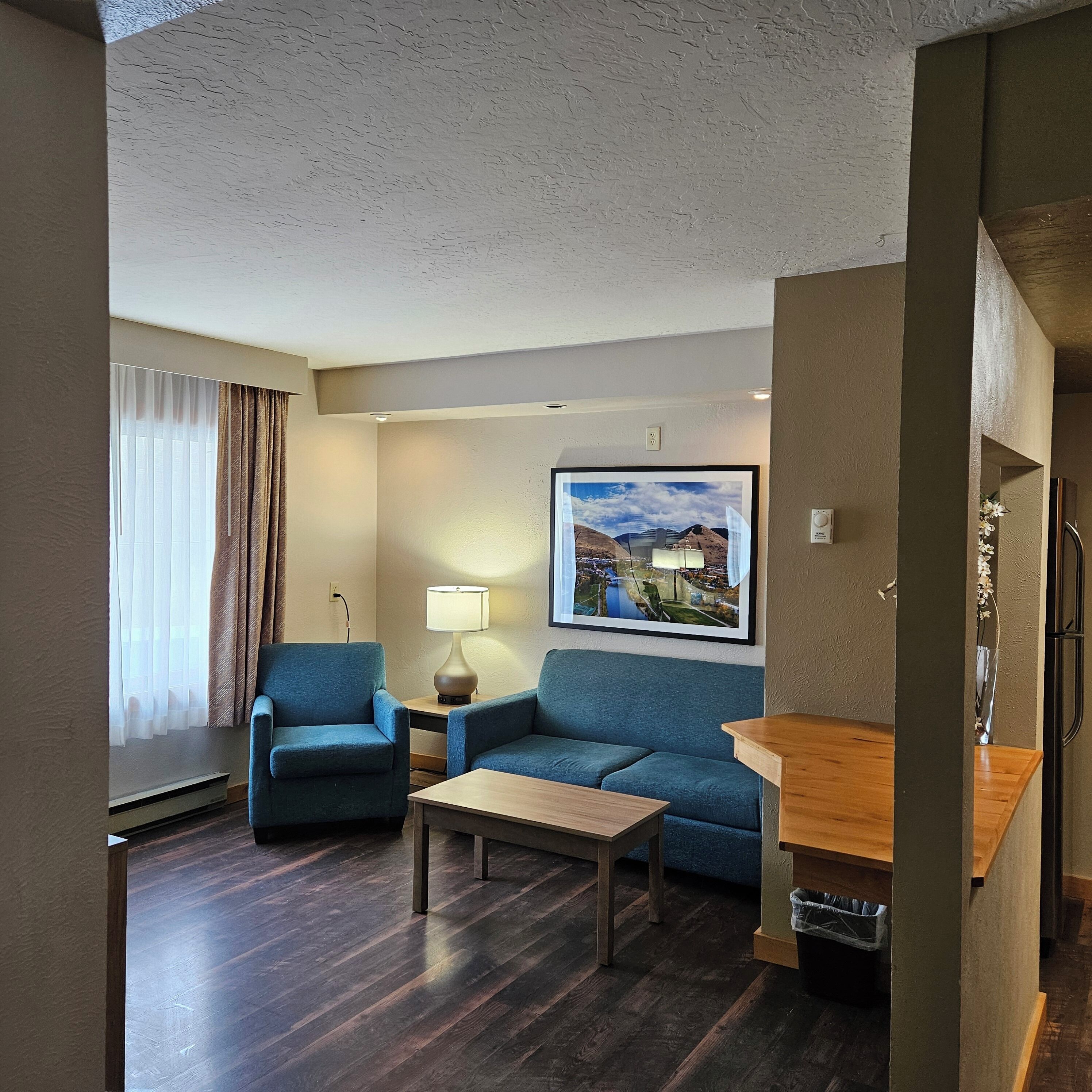 A hotel room with a blue sofa, a wooden coffee table, a lamp, and a painting of a landscape.