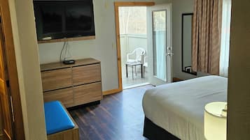 A hotel room with a bed, a TV mounted on the wall, a wooden dresser, a blue bench, and a sliding glass door leading to an outdoor area with chairs.