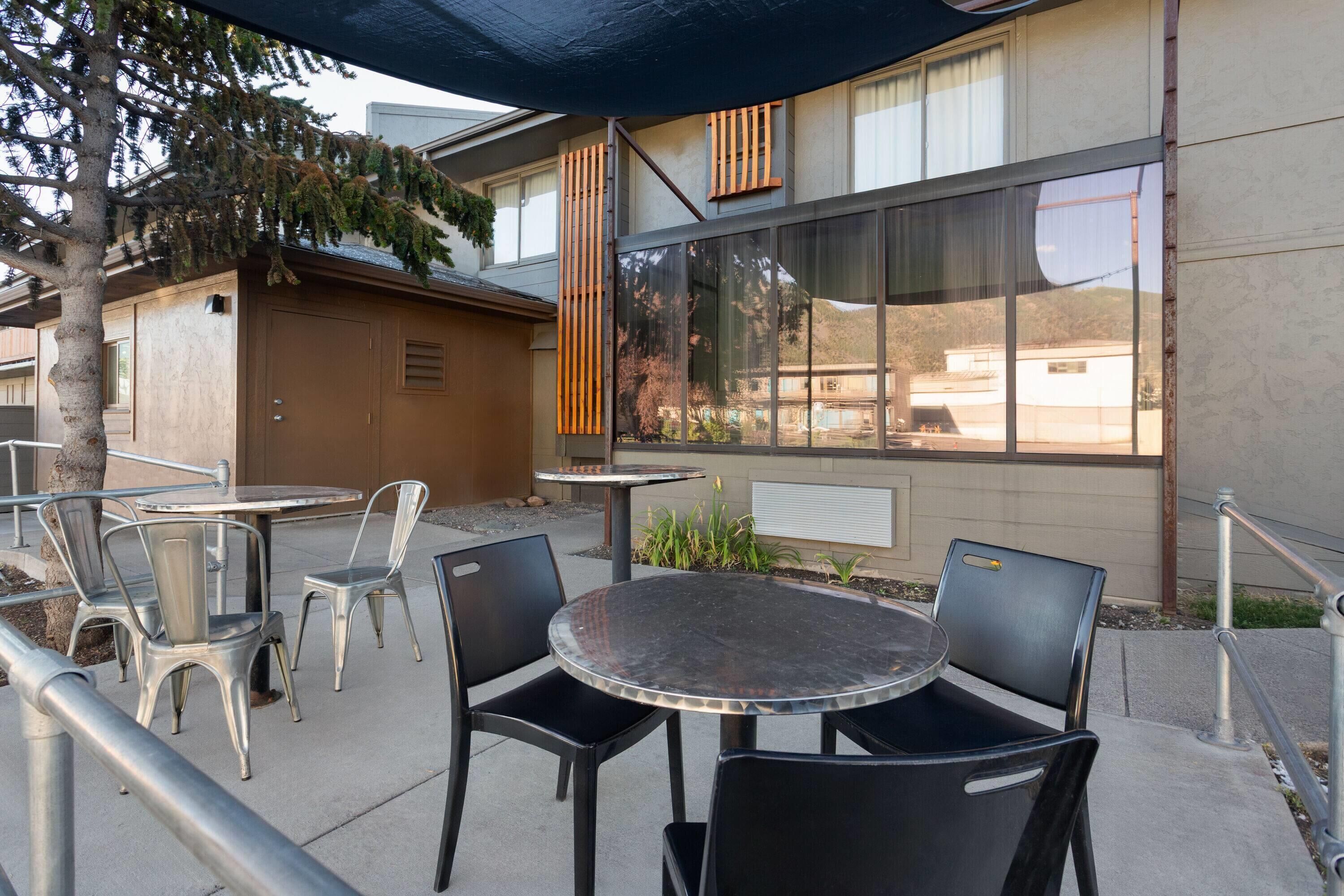 Une terrasse extérieure avec des tables et des chaises rondes, un bâtiment doté de grandes fenêtres et un arbre.