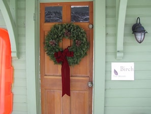 A wooden door with a red bow, a green wreath with pine cones, and a sign reading "Birch The Woods".