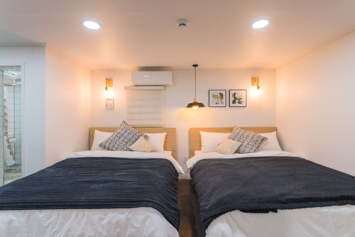 A hotel room with two beds, a ceiling fan, wall-mounted air conditioning, and a small window.