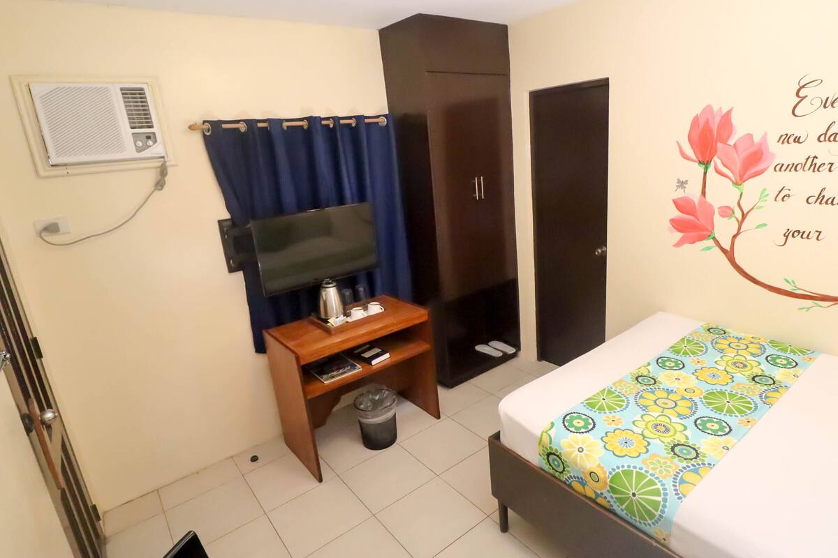 A hotel room with a bed, a TV, a wardrobe, a small table with a kettle, and a trash bin.