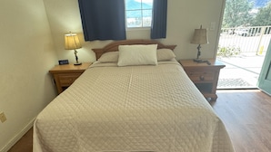 A bedroom with a large bed, wooden headboard, two bedside tables with lamps, a window with curtains, and a view of the outdoors.