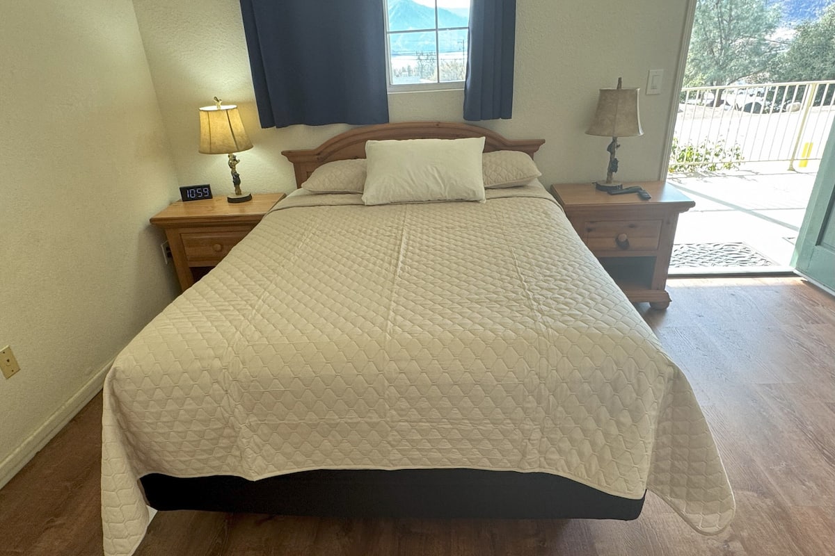 Une chambre avec un grand lit, une tête de lit en bois, deux tables de chevet avec des lampes, une fenêtre avec des rideaux et une vue sur l’extérieur.