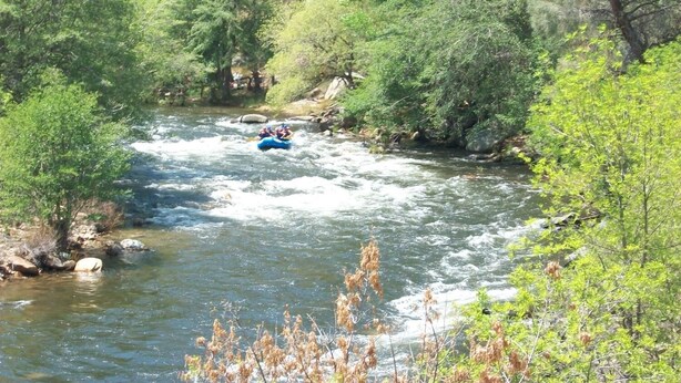 A river with a blue raft navigating through it, surrounded by dense green foliage. - Lakeshore Lodge (Wofford Heights)