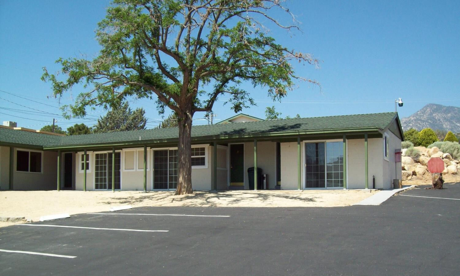 A motel with a large tree in front, a parking lot, and a mountain in the background.