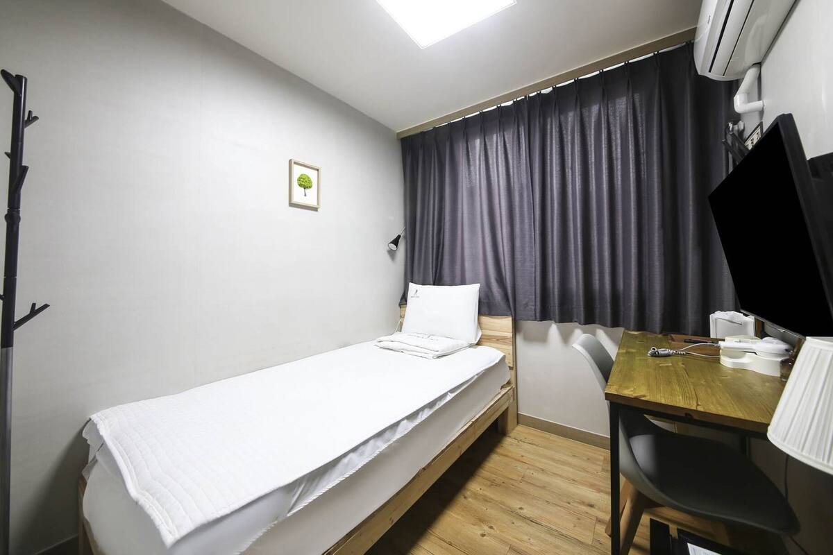 Une petite chambre avec un lit simple, un plancher en bois, un bureau avec une chaise, une télévision et une fenêtre avec des rideaux.