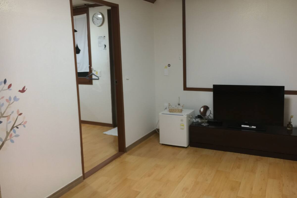 Une pièce avec un plancher en bois, un téléviseur posé sur un meuble, un petit réfrigérateur et une porte donnant sur une autre pièce.