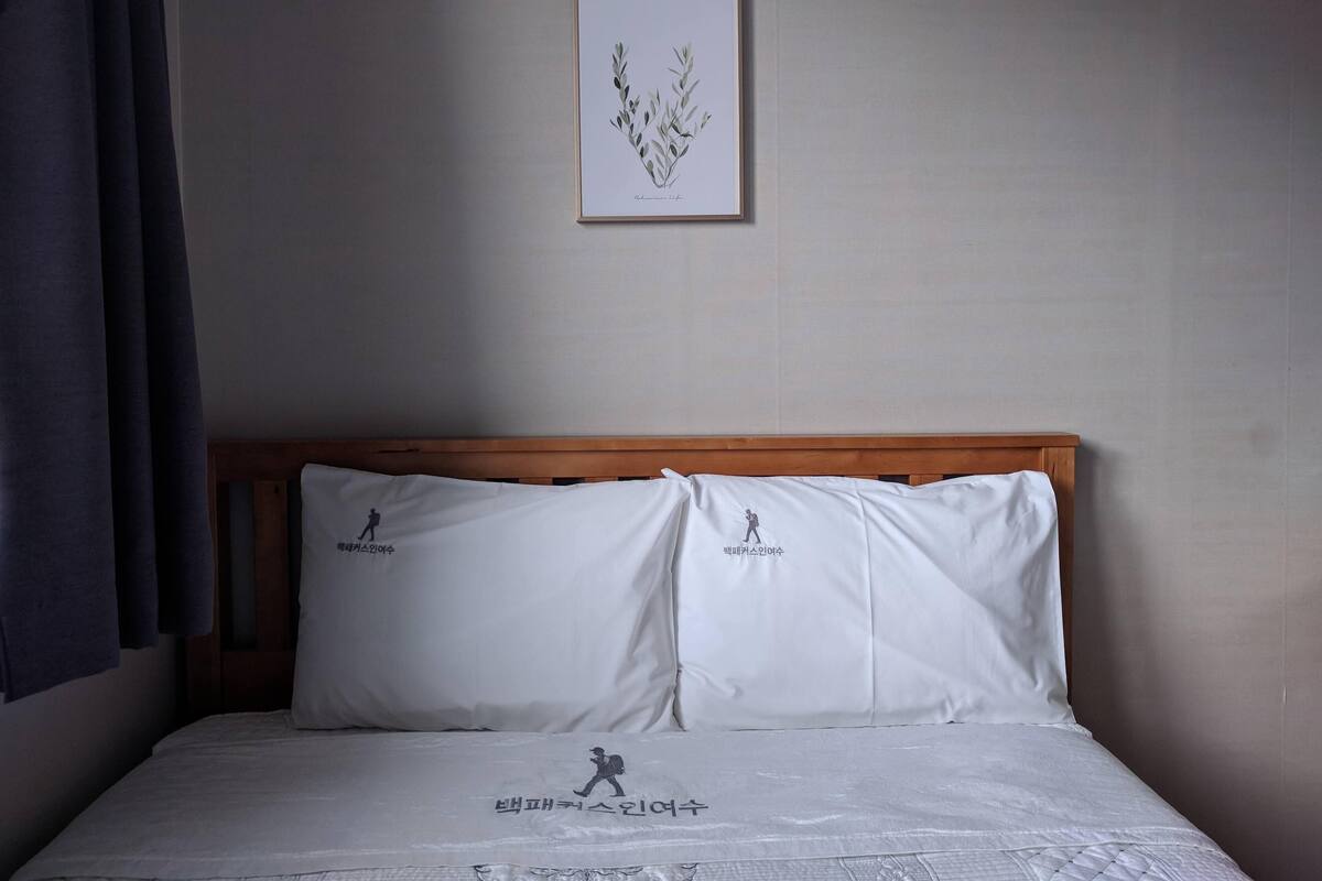 Un lit bien fait, avec une literie blanche et deux oreillers ornés d’une silhouette de coureur. Une estampe botanique encadrée est accrochée au mur, au-dessus du lit.