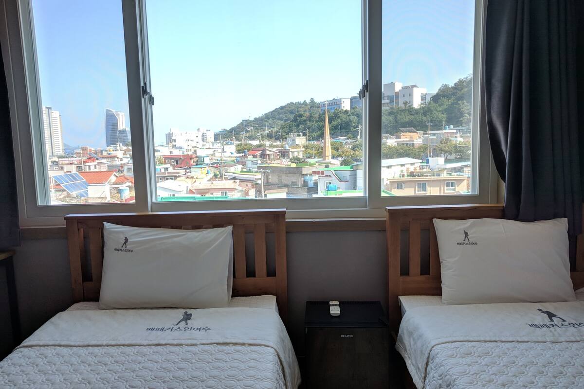 Une chambre d’hôtel avec deux lits, une vue sur le paysage urbain grâce à de grandes fenêtres, et une petite table placée entre les lits.