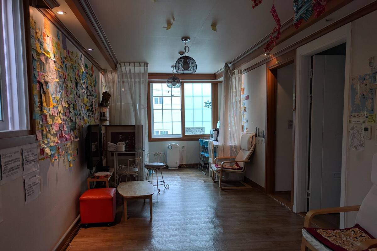 Une pièce avec un plancher en bois, un pouf rouge, une chaise, une petite table et une fenêtre avec des rideaux.