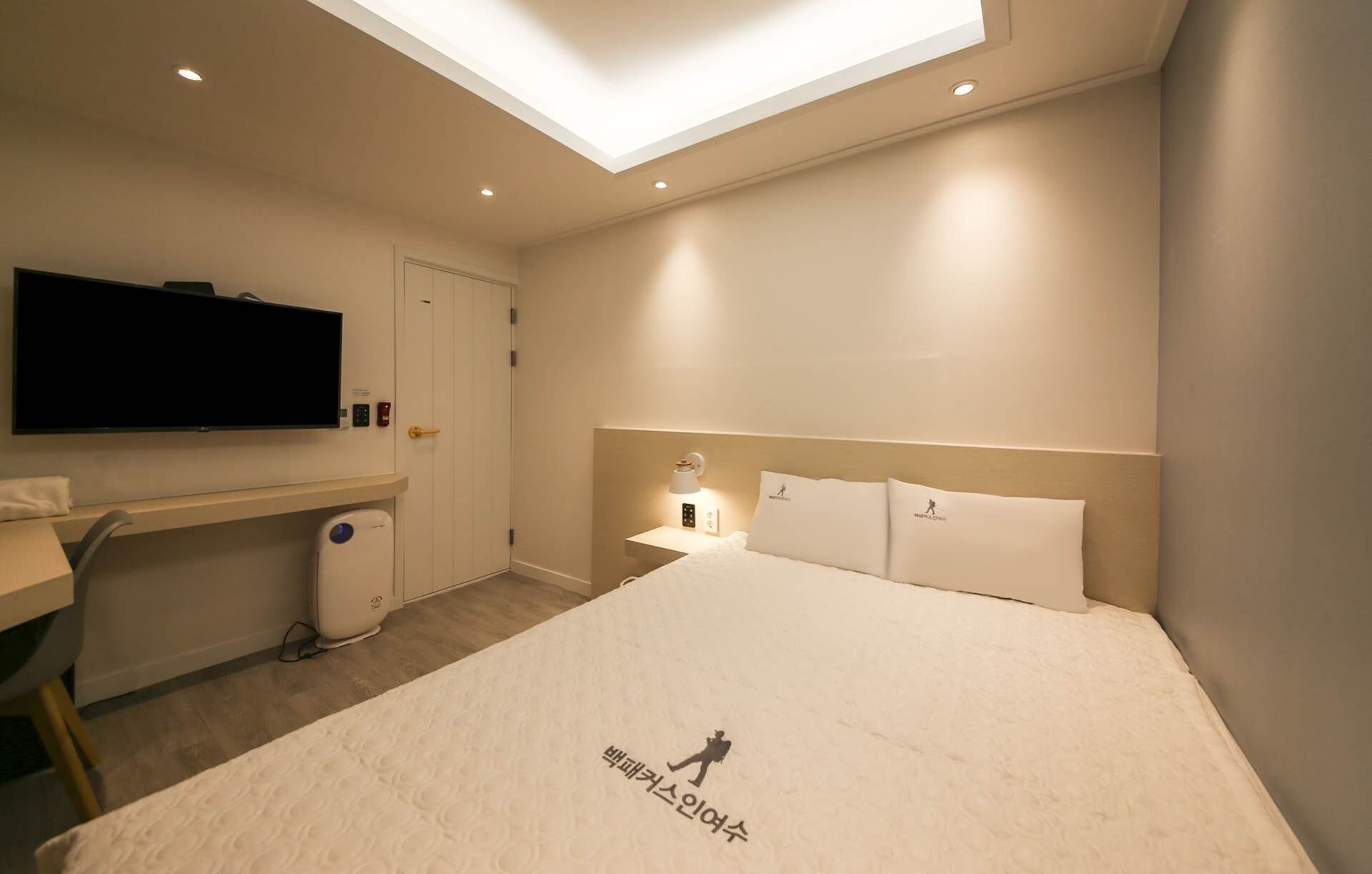 A hotel room with a large bed, a desk with a flat-screen TV, a bedside table with a lamp, and a ceiling light.