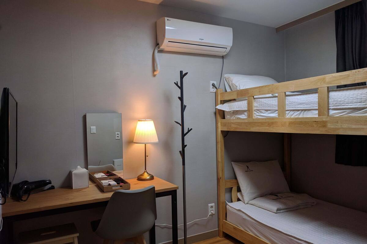 Une chambre d’hôtel compacte comprenant un lit superposé, un bureau avec une lampe, une chaise, un miroir et un appareil de climatisation.