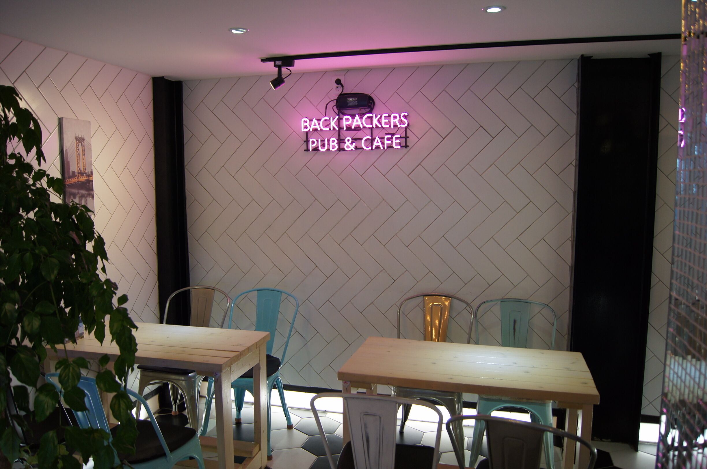 A modern interior with a herringbone-patterned wall, a neon sign reading "BACK PACKERS PUB & CAFE", and a seating area with metal chairs and wooden tables.