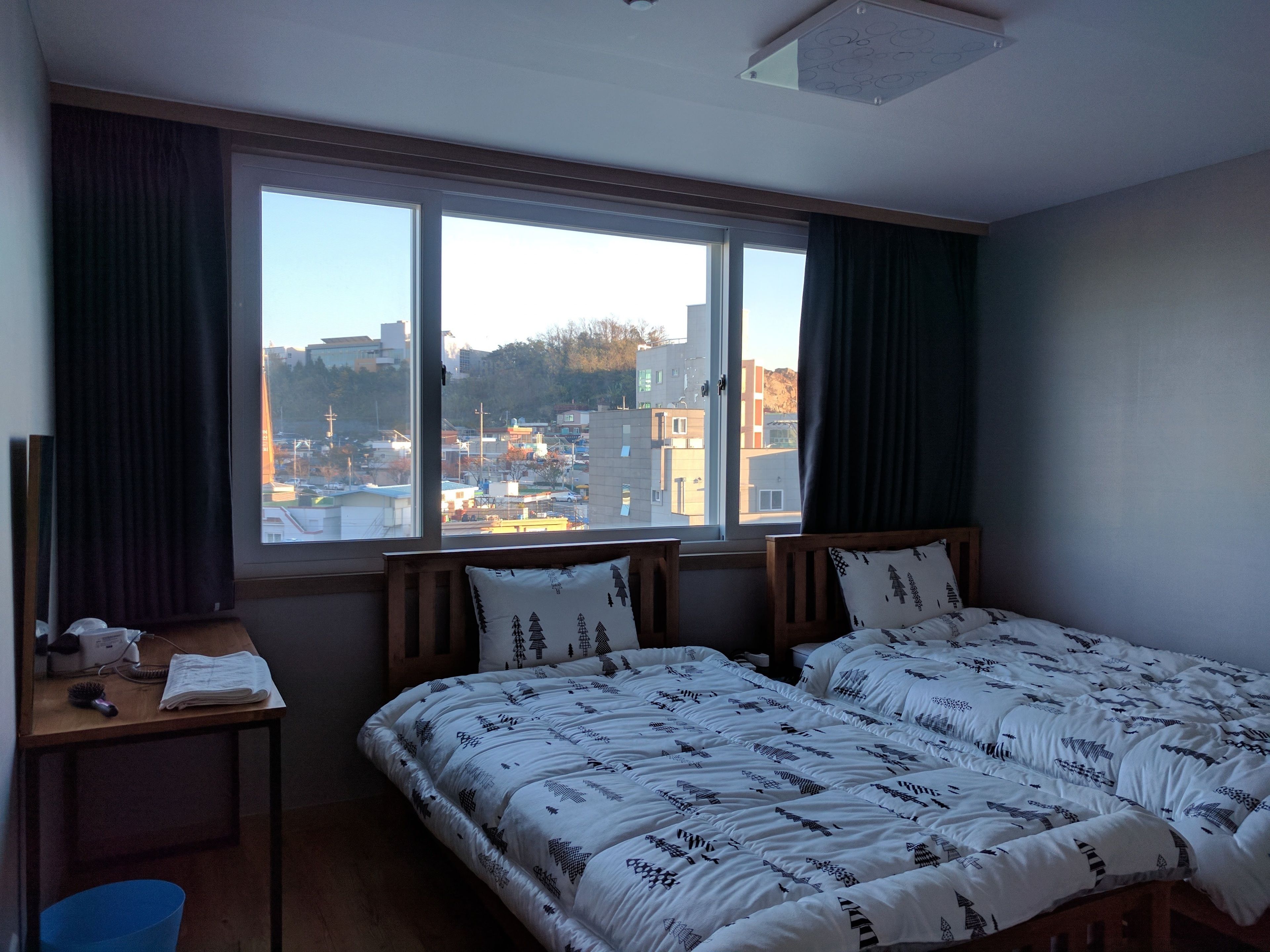 A hotel room with two beds, a desk, and a large window offering a city view.