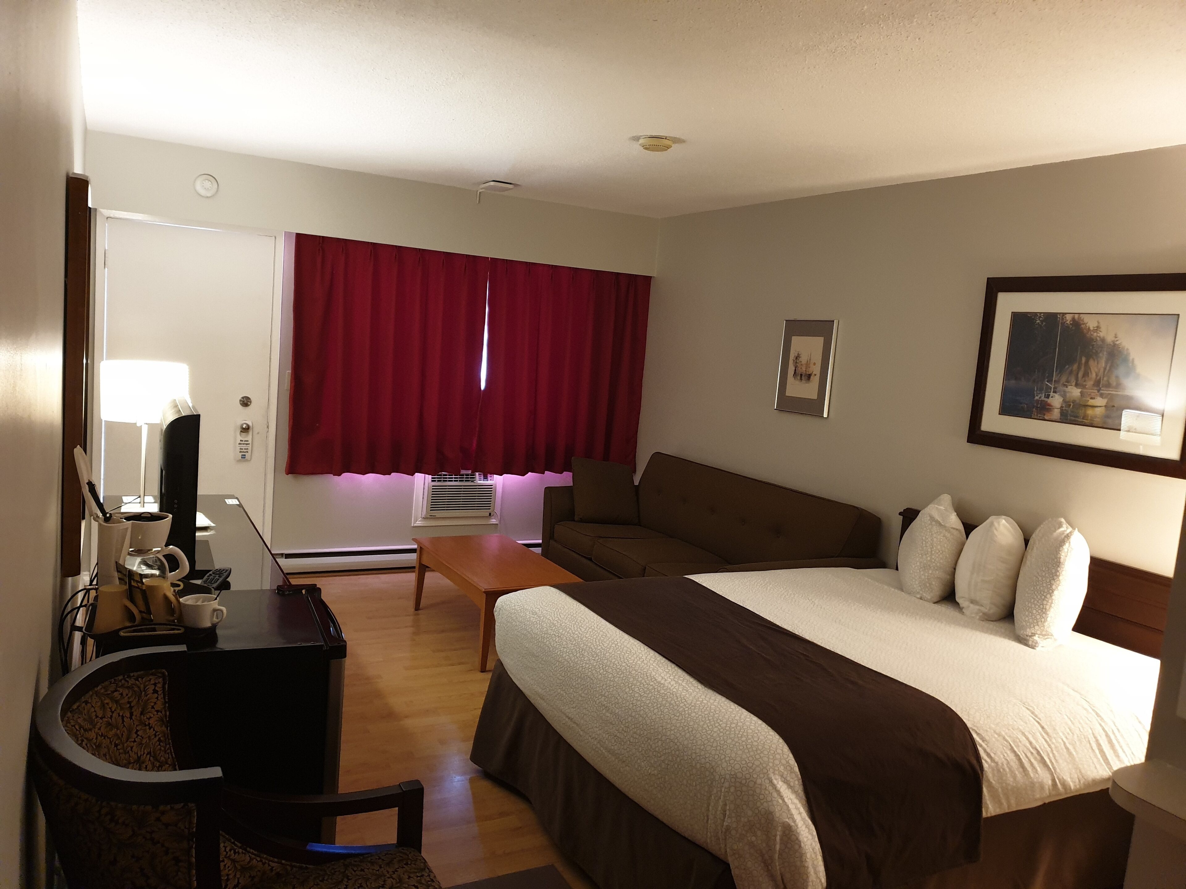 A hotel room with a bed, a desk with a lamp, a chair, a sofa, and a window with red curtains.