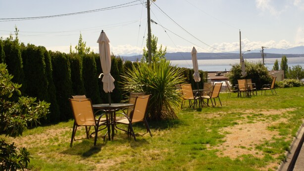 An outdoor seating area with tables and chairs, surrounded by greenery and overlooking a body of water with mountains in the distance. - Island View Lodge (Powell River)