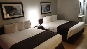 A hotel room with two beds, each with white bedding and dark headboards. There are framed pictures on the walls, a bedside table with a lamp, and a window with a view of the outside. - Island View Lodge (Powell River)