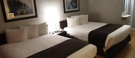 Une chambre d’hôtel avec deux lits, chacun doté de draps blancs et d’une tête de lit sombre. Les murs sont ornés de tableaux encadrés, il y a une table de chevet avec une lampe, et une fenêtre donnant sur l’extérieur.