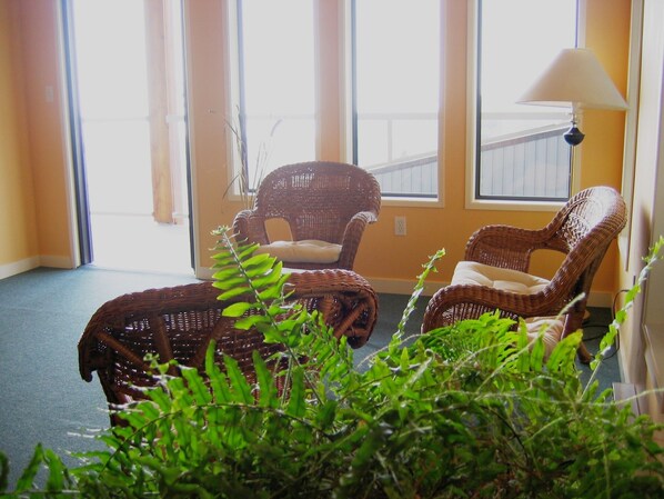 Una habitación con dos sillas de mimbre y una mesita, ventanas amplias con persianas y una lámpara.