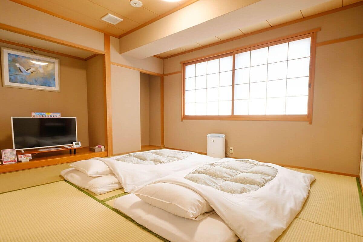 A traditional Japanese room with tatami flooring, a futon bed, a television, and a window with shoji screens.
