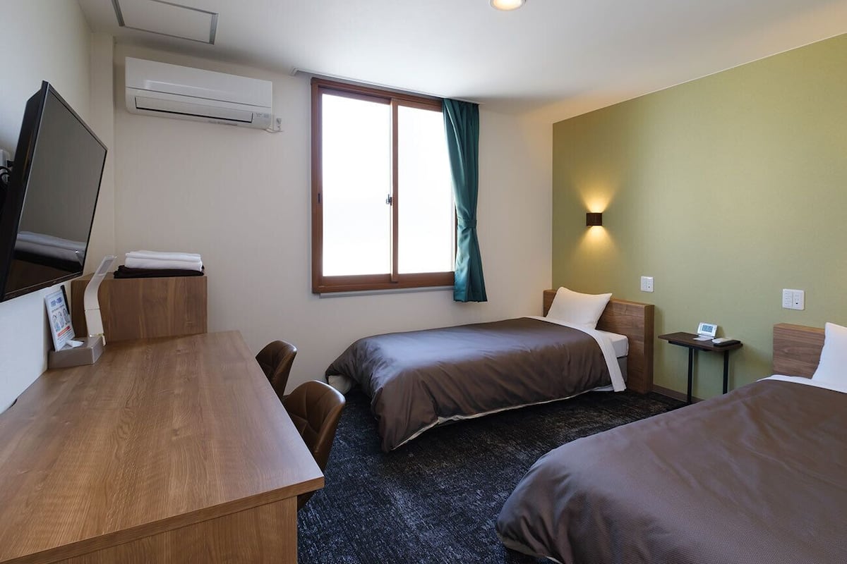 A hotel room with two beds, a desk, a TV, and a window with curtains.